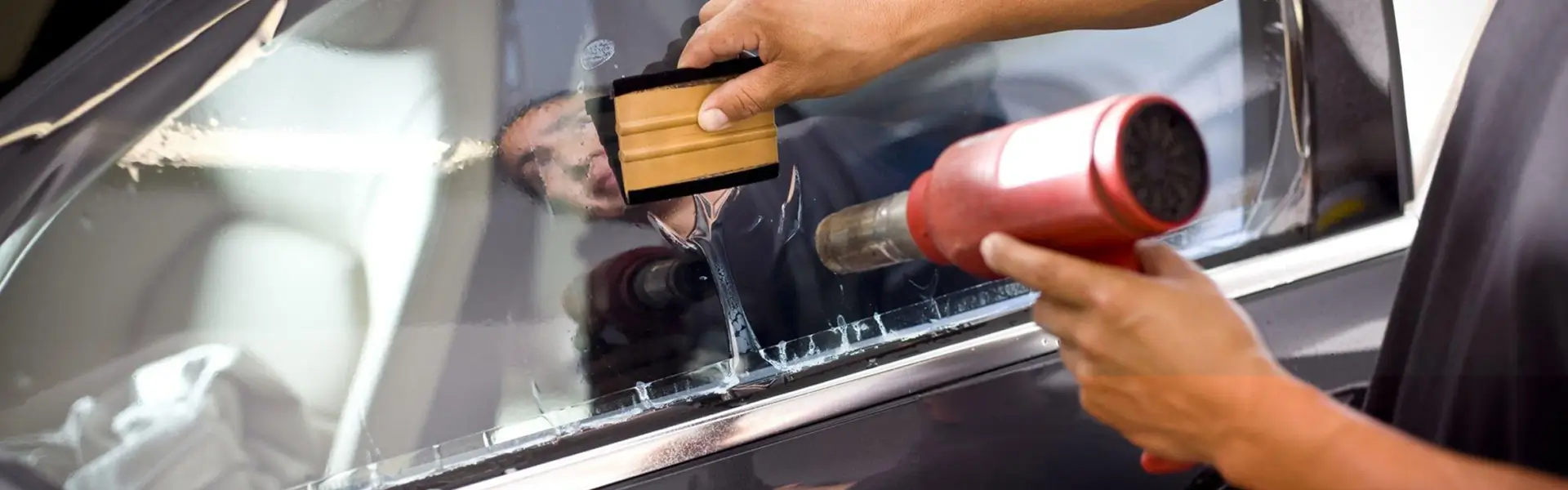 Close-up of a professional installing automotive window tint using a heat gun and squeegee tool on a side window, carefully smoothing the film for a flawless finish.