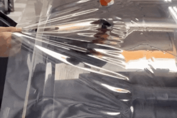 A person applies a clear, flexible sheet of window tint film onto a car window, smoothing it out by hand to ensure a proper fit. The film creates a protective layer that controls sunlight and UV exposure inside the vehicle.