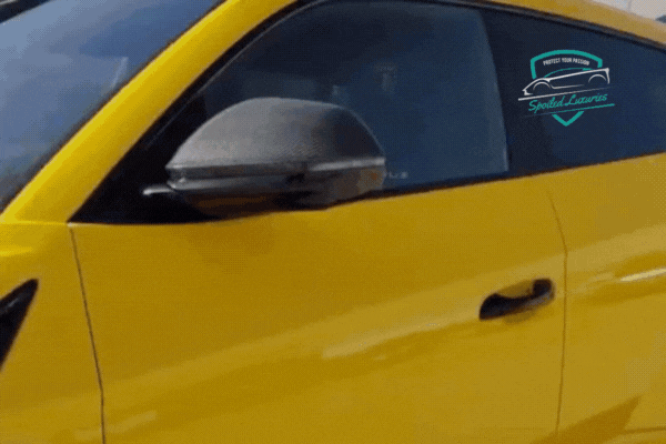 A close-up of a yellow car’s side window as a technician professionally installs window tint, ensuring a smooth, bubble-free finish. The Spoiled Luxuries logo is visible on the window.
