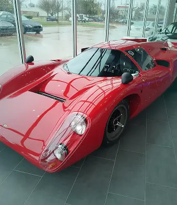A bright red classic race car with sleek curves and a low profile is parked inside a glass-walled showroom on a rainy day, showcasing its glossy, pristine finish.