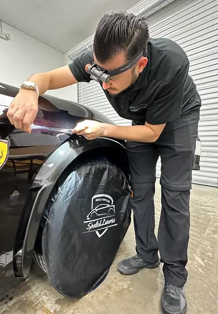 An auto detailer wearing a headlamp and black uniform uses a precision tool to inspect or correct the paint near the fender of a black Ferrari. The front wheel is covered with a branded “Splash Coater” wheel protector.