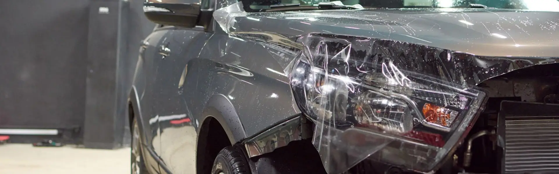 A clear paint protection film is partially applied to the front end of a wet, dark gray SUV, with the film draped over the headlight and hood area during installation.