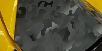 Close-up of a vehicle hood wrapped in a matte black and gray camouflage vinyl, bordered by vibrant yellow paint on the fenders.
