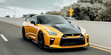 A yellow and black Nissan GT-R sports car driving on a highway, featuring an aggressive front design and custom black hood, with trees and buildings in the background.