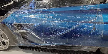 Close-up of a blue sports car with a transparent paint protection film (PPF) being applied, showing water bubbles and alignment on the body panel.