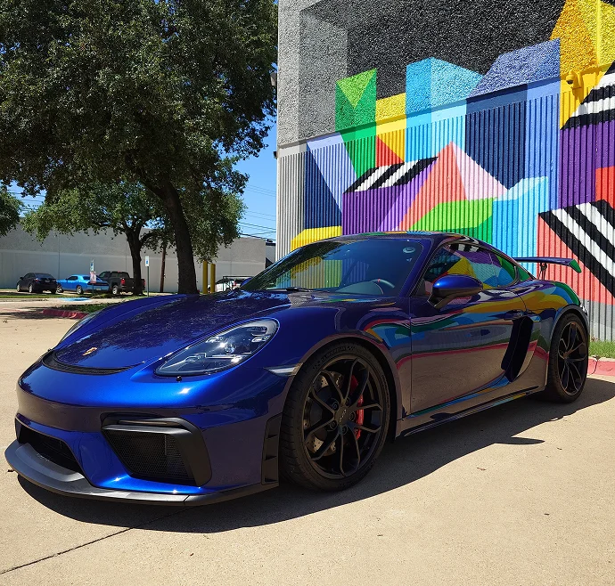 A deep blue Porsche 718 Cayman GT4 with black wheels and red brake calipers is parked on a sunlit pavement in front of a colorful geometric mural.
