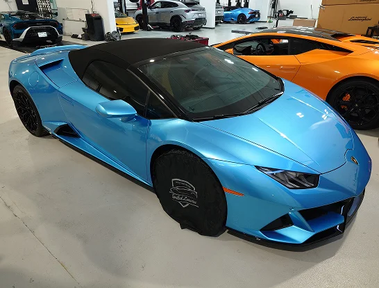 Sky blue Lamborghini Huracán Spyder with a black convertible top is parked indoors, featuring protective wheel covers branded “Splash Coater.” Nearby are other exotic cars including an orange Lamborghini.