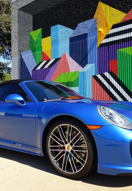Front-side view of a blue Porsche 911 Carrera parked in front of a vibrant geometric mural, highlighting the car's detailed alloy wheels and glossy paint finish.