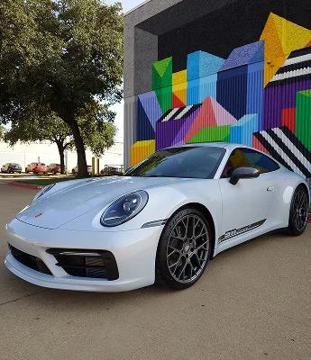 A white Porsche 911 with black trim and dark alloy wheels is parked in front of a colorful geometric mural, showcasing its clean lines and freshly detailed finish.