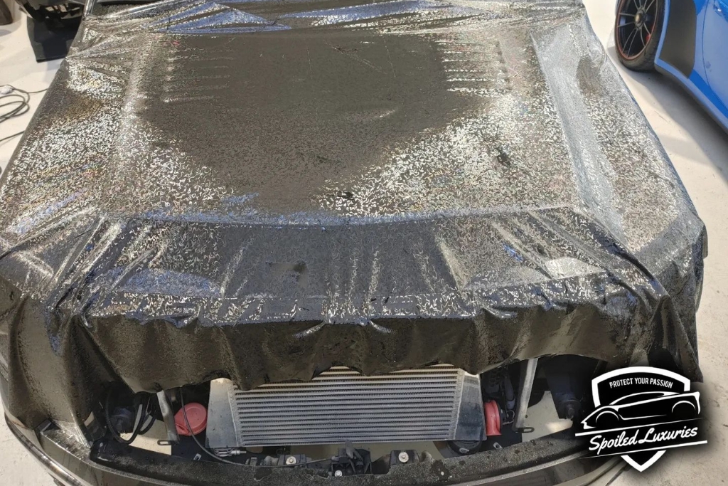 Close-up of a car hood partially wrapped in a glittery black vinyl film during the application process at Spoiled Luxuries. The film is still being positioned and smoothed out, showing an early stage of how vinyl wrapping works.