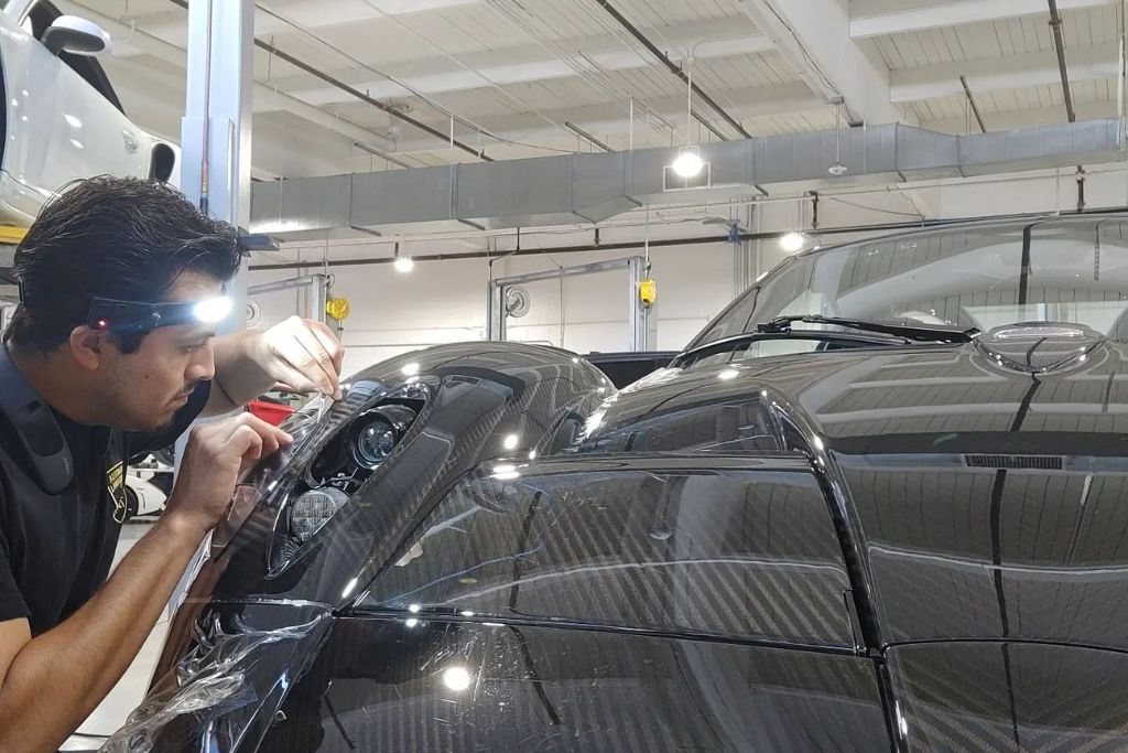 A technician wearing a headlamp carefully applies Paint Protection Film around the headlight of a black carbon-fiber supercar inside a well-lit garage. This meticulous process illustrates the professional installation that impacts the answer to How Long Does PPF Last.