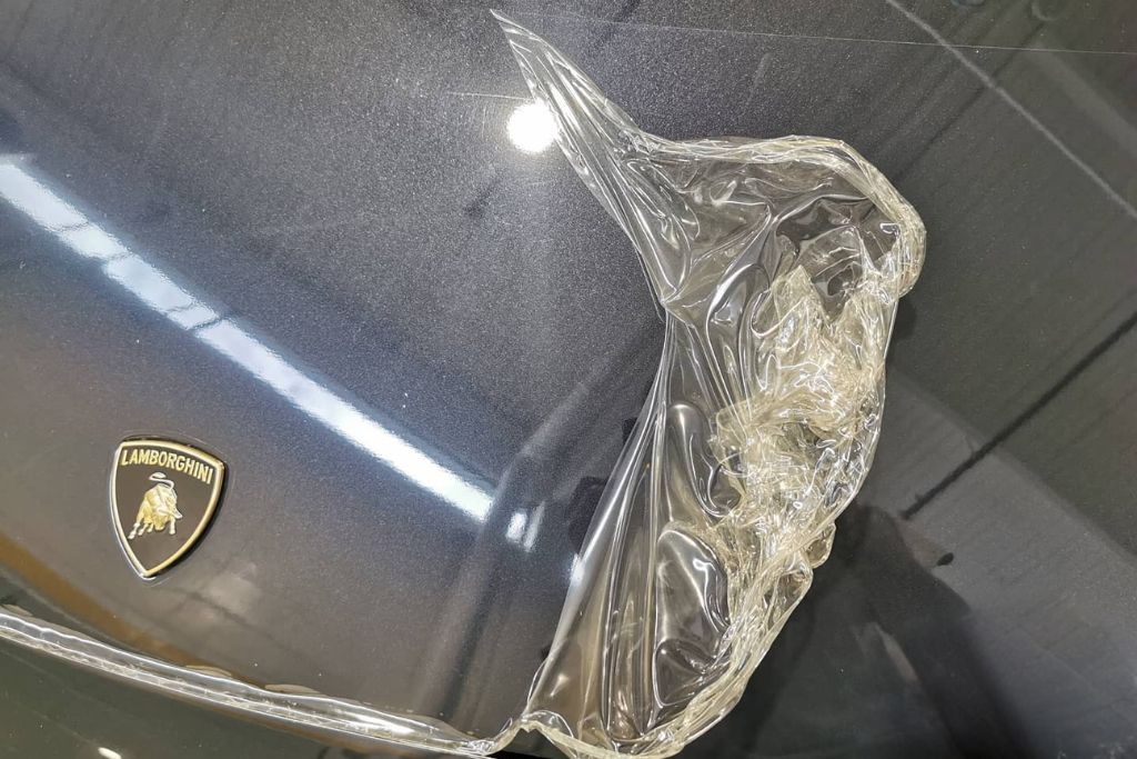 PPF stands for Paint Protection Film, a nearly invisible, high-performance layer of thermoplastic urethane film that is professionally applied to a vehicle's exterior surfaces. Imagine it as a shield, similar to a screen protector for your phone, but engineered to withstand far harsher conditions. Originally developed by the U.S. military during the Vietnam War to protect helicopter rotor blades from flying debris, the technology has since evolved for civilian automotive use. Today's PPF is a sophisticated, multi-layered material designed to absorb impacts, resist scratches, and block harmful UV rays, all while maintaining your car's natural shine. Close-up of a clear Paint Protection Film (PPF) being peeled off the glossy black hood of a Lamborghini, revealing a pristine paint surface beneath. This visual demonstrates how PPF acts as an invisible shield against damage, answering the question “What is PPF” in the context of automotive care.