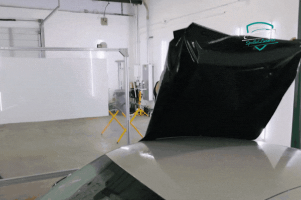 Vinyl wrap being precisely applied to the roof of a car inside a clean, climate-controlled garage at Spoiled Luxuries in Dallas. The black film is being aligned and positioned before smoothing, highlighting professional installation standards.