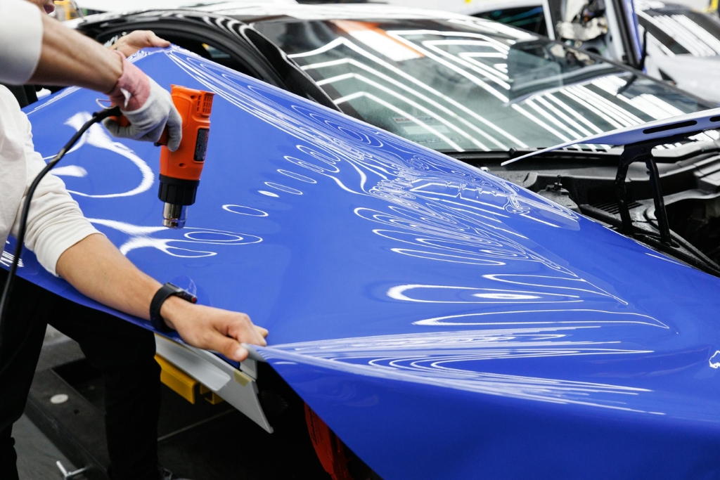 Installer uses a heat gun to apply and mold a glossy blue vinyl wrap onto a car hood, with light reflections emphasizing the wrap's smooth finish. This demonstrates a key stage in choosing vinyl wrapping over traditional paint for both speed and style.