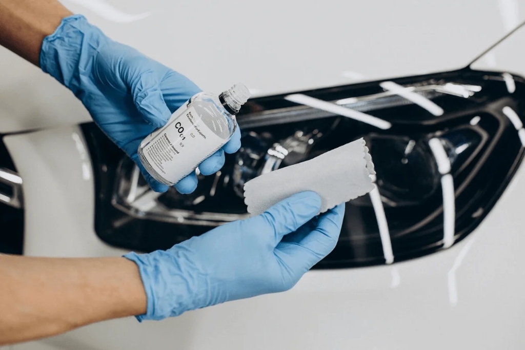 A person wearing blue gloves holds a ceramic coating bottle labeled “CO01” and a suede applicator cloth in front of a clean white car headlight, preparing for precise application.