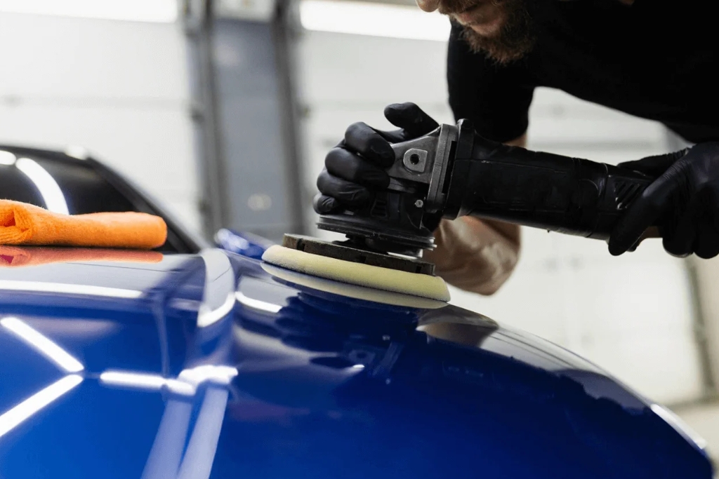 A professional uses a dual-action polisher on the glossy blue hood of a car, preparing the surface for ceramic coating. An orange microfiber towel rests nearby, and the reflection of shop lighting highlights the polished finish.