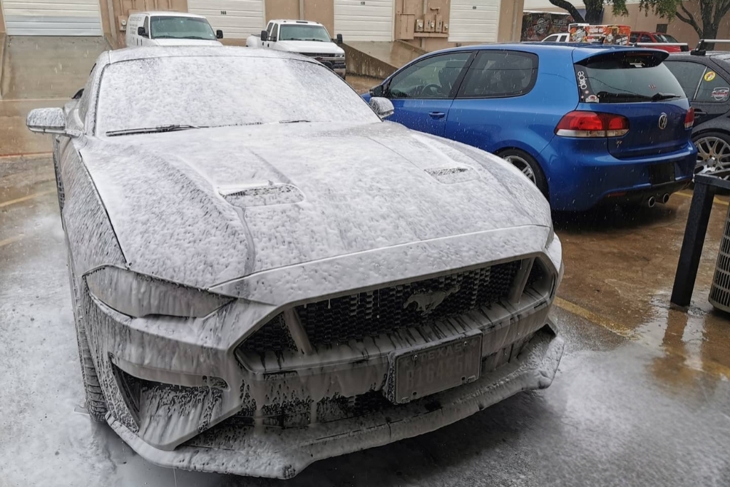 A Ford Mustang is completely covered in thick white foam during a pre-wash outside a detailing shop, with a blue Volkswagen parked nearby. The foam wash is part of maintaining a ceramic coating without damaging it.