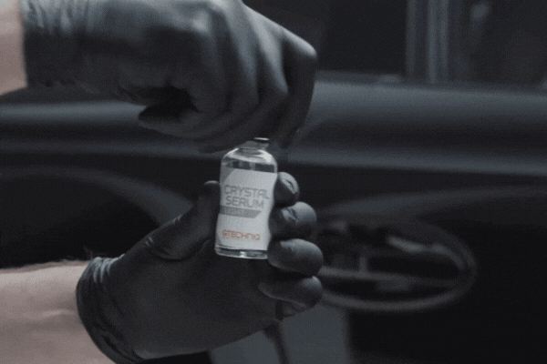 A person wearing black gloves opens a small bottle of Gtechniq Crystal Serum Light ceramic coating in front of a car door, preparing for application. The movement highlights the careful handling required for ceramic coating.