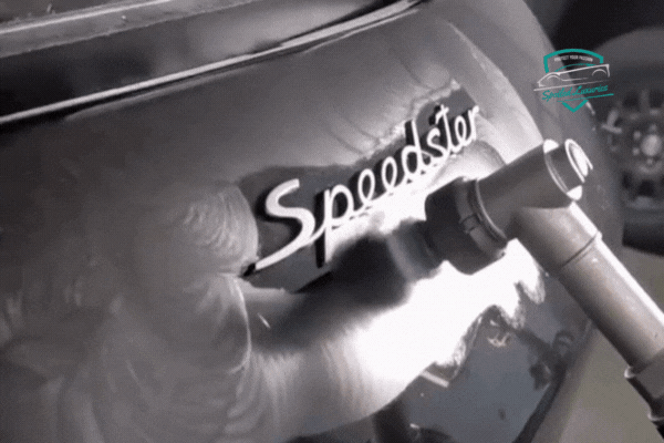 A detailer wearing gloves uses a small inspection light to check the ceramic-coated surface near the Speedster badge on a dark-colored Porsche. The paint reflects light clearly, showcasing the coating's high gloss and smoothness. Spoiled Luxuries logo appears in the top right.