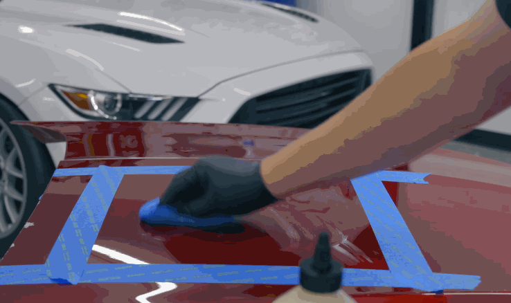 A gloved hand spreads ceramic coating evenly over a taped-off section of a red car hood using an applicator pad.