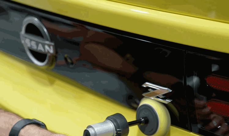 A power tool with a polishing pad is used to refine and buff the rear emblem of a yellow Nissan Z, likely before applying wax or ceramic coating.