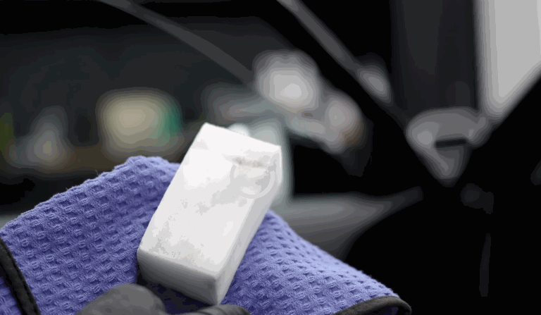 Close-up of a hand holding a white magic eraser sponge above a folded purple microfiber towel, tools often used in car detailing.