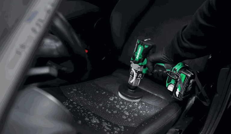 A gloved hand operates a cordless drill brush on a soapy fabric car seat to lift dirt during interior car detailing.