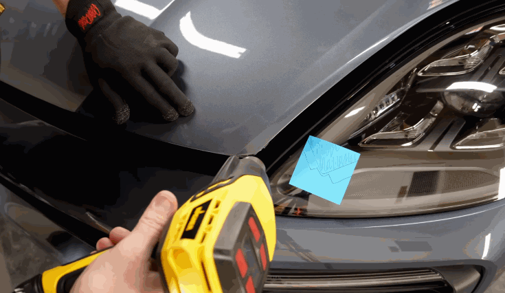 Close-up of a person using a heat gun to apply paint protection film to a car's headlight area, while another person holds the hood steady with gloved hands.