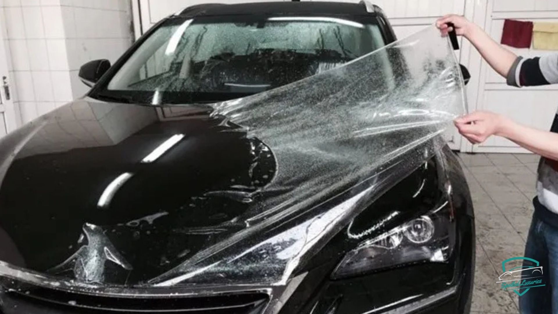 Person applying paint protection film to the hood of a black car in a garage, with the transparent film partially peeled back and water droplets visible underneath.