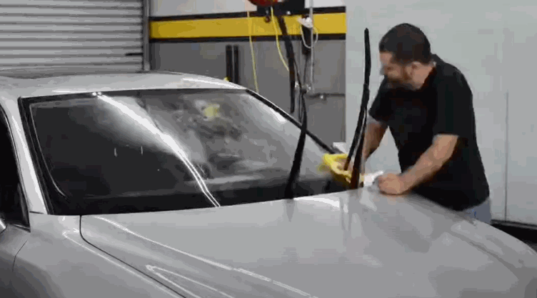 Man cleaning the windshield of a silver car in preparation for window tinting, with the wipers lifted for access.