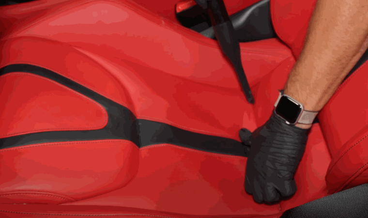 Close up of a red leather car seat being cleaned by a gloved hand during car detailing. The seat surface appears smooth and well maintained as part of an interior restoration process.