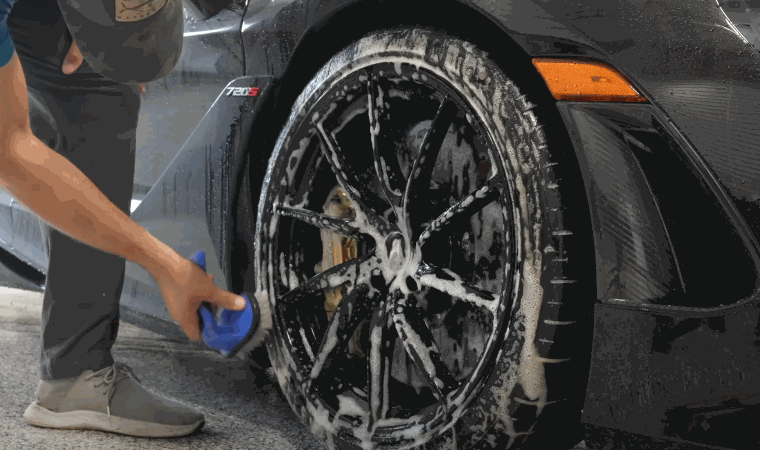 Person scrubbing a black alloy wheel with a blue brush during car detailing. Thick white soap covers the tire and rim to remove brake dust and road grime.